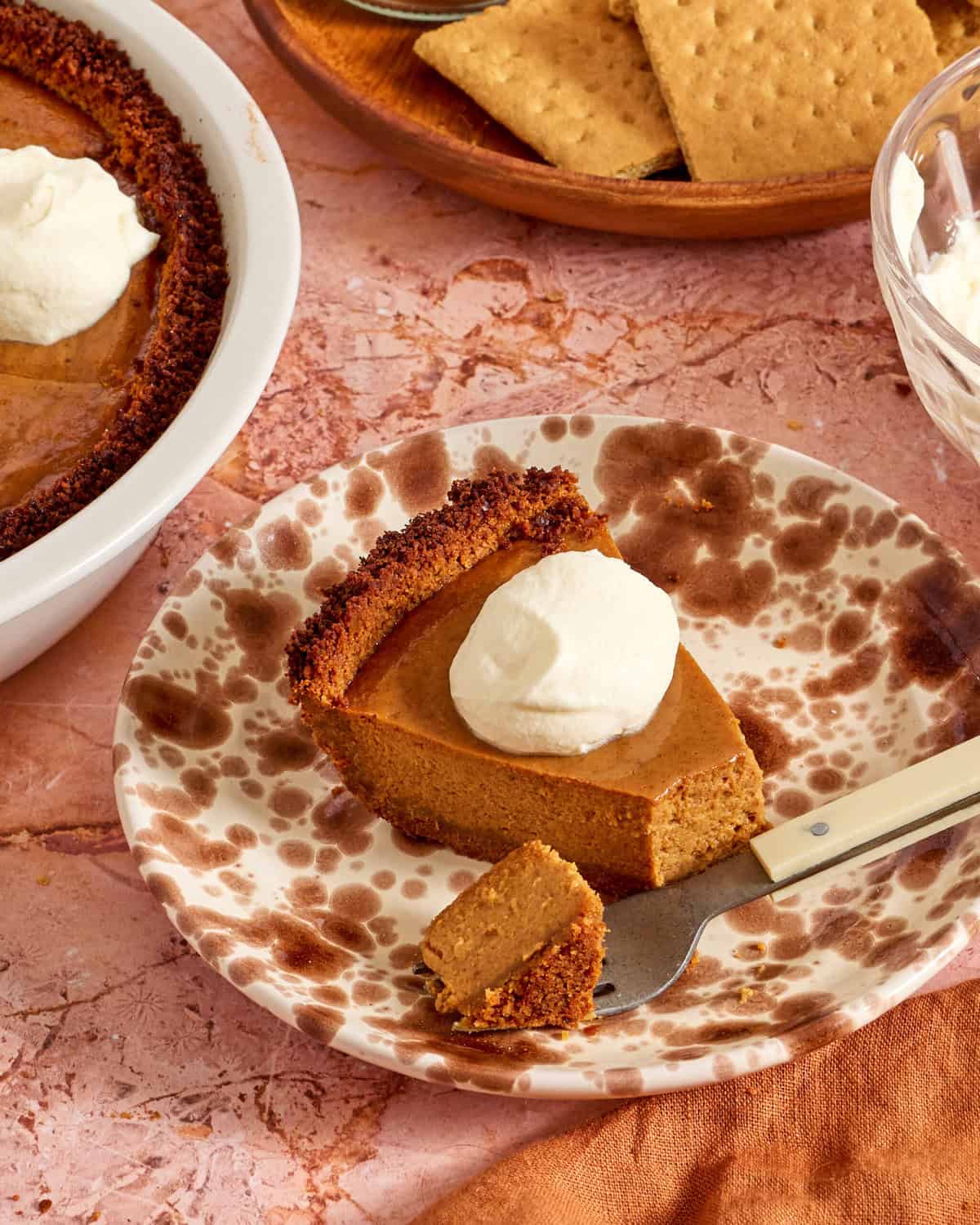 A slice of pumpkin pie with a graham cracker crust topped with whipped cream sits on a plate. A fork holds a small bite. In the background, the rest of the pie, more graham crackers, and a bowl of whipped cream are visible.