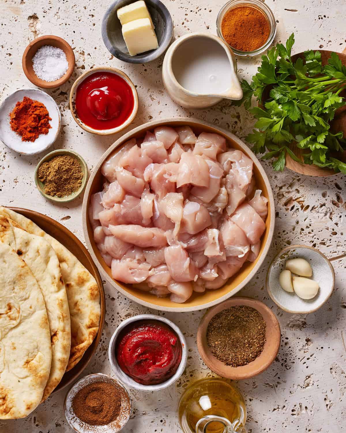 Overhead view of raw diced chicken in a bowl surrounded by naan bread, various spices, garlic cloves, butter, ketchup, tomato paste, fresh herbs, and a small pitcher of cream on a light textured surface.