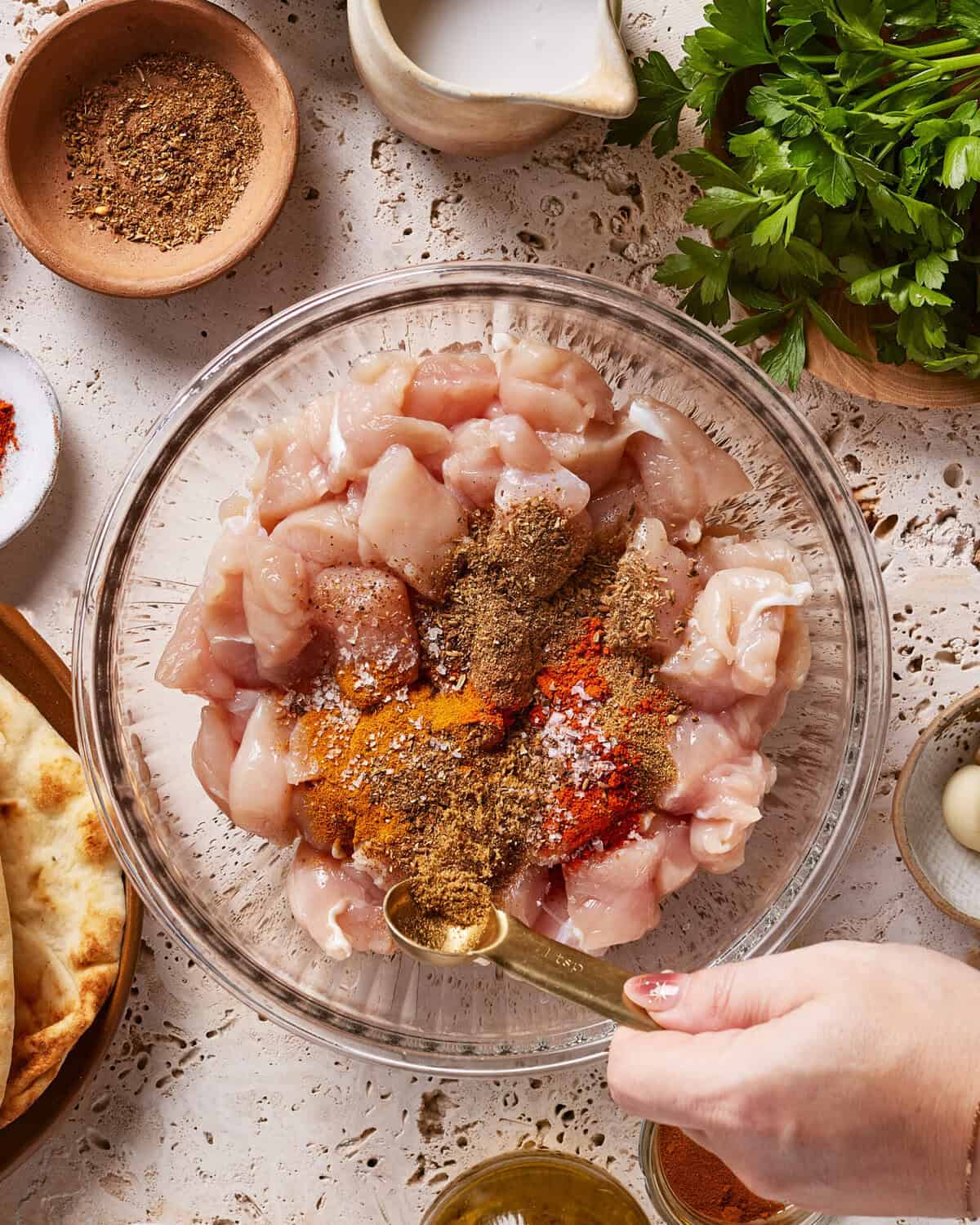 A hand adds ground spices to a glass bowl filled with diced raw chicken. Surrounding the bowl are fresh parsley, naan bread, olive oil, spice bowls, a garlic clove, and a small jug of cream on a lacy tablecloth.