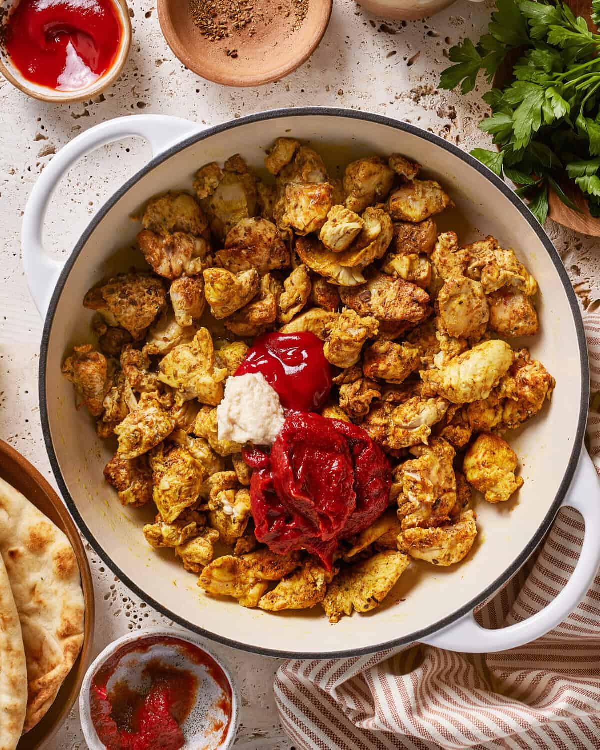 A white pot filled with seasoned, cooked chicken pieces topped with ketchup and tomato paste, surrounded by naan bread, ketchup, parsley, spices, and a striped cloth on a light table.