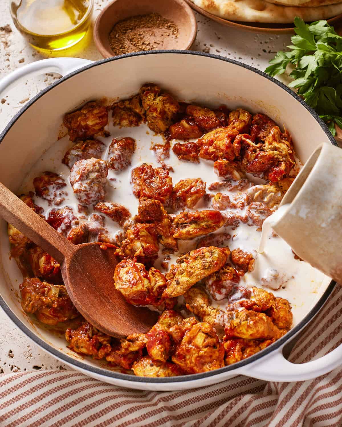 A pot of spiced chicken pieces cooked in a tomato sauce, with cream being poured in, and a wooden spoon resting inside. Fresh herbs, spices, olive oil, and naan bread are nearby on the table.