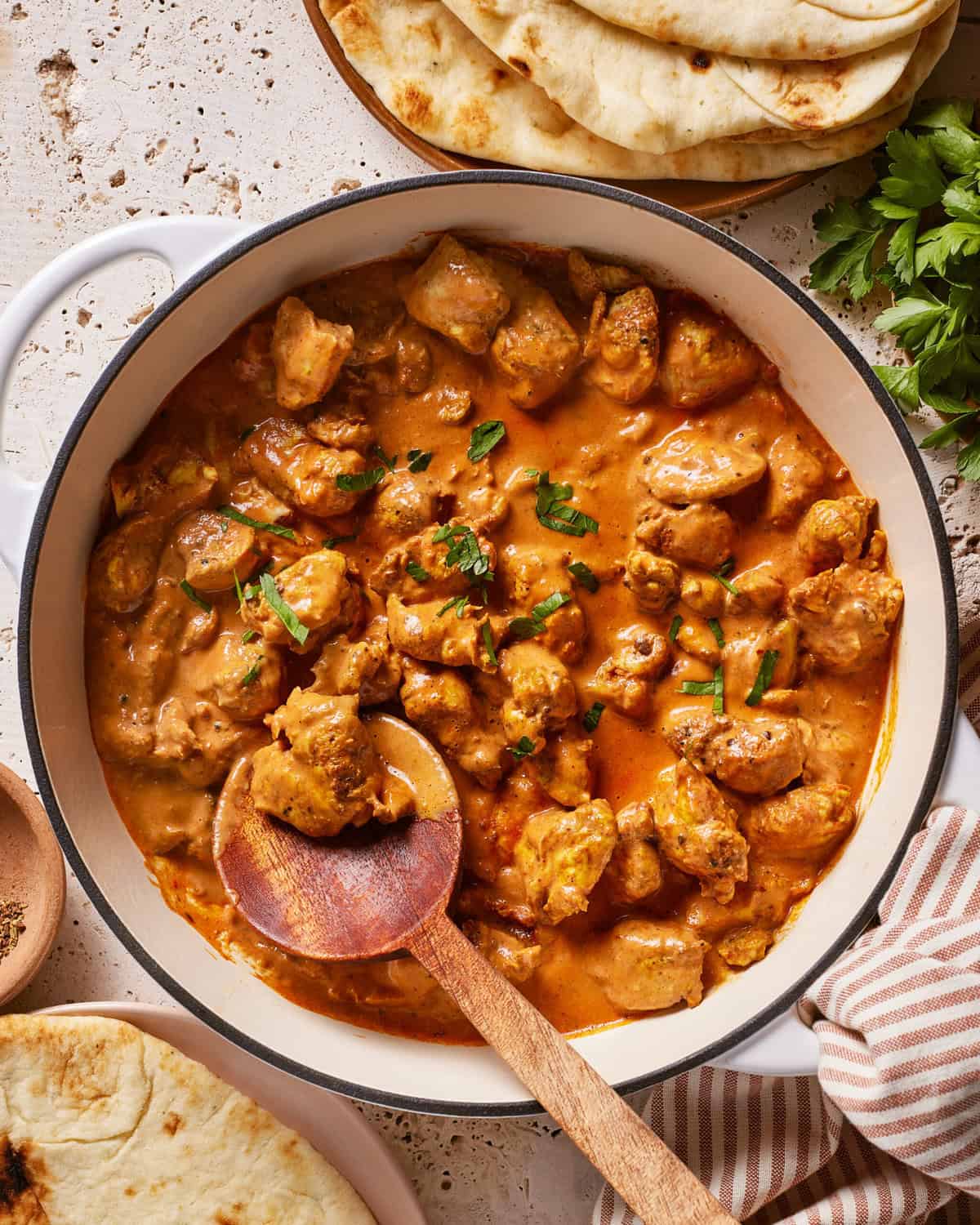 A white pot filled with creamy, orange-brown chicken curry garnished with fresh herbs, with a wooden spoon resting inside. Naan bread and fresh parsley are visible nearby on a textured surface.