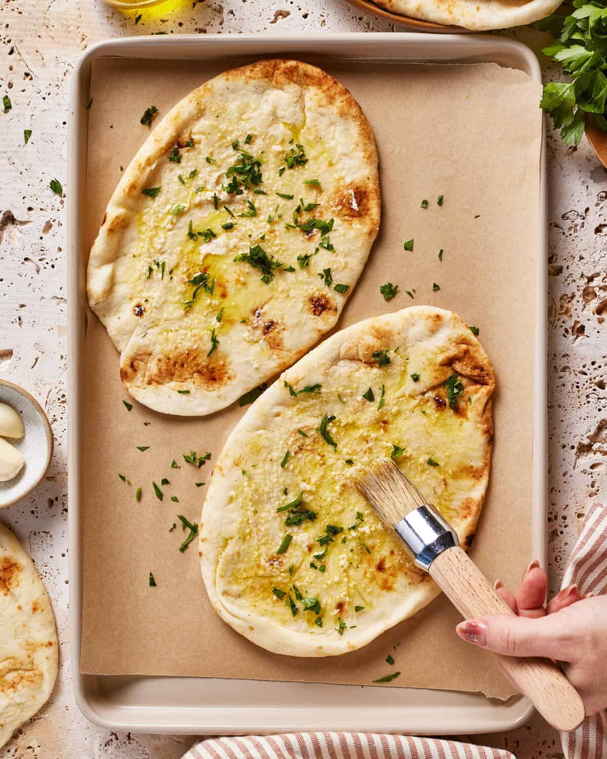 Two pieces of flatbread on parchment paper are being brushed with garlic oil and sprinkled with chopped parsley. A hand holds a pastry brush, and fresh herbs and garlic cloves are nearby.