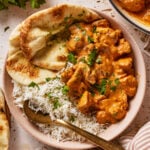 A bowl of butter chicken in creamy orange sauce, served with white basmati rice, naan bread, and garnished with fresh chopped parsley. A gold spoon rests next to the rice.
