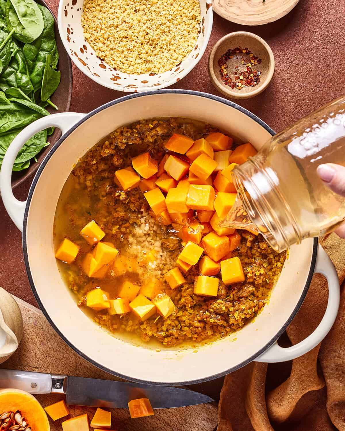 A hand pours liquid from a glass jar into a pot filled with chopped butternut squash, onions, and spices. Nearby are bowls of spinach, millet, red pepper flakes, and a knife on a cutting board.