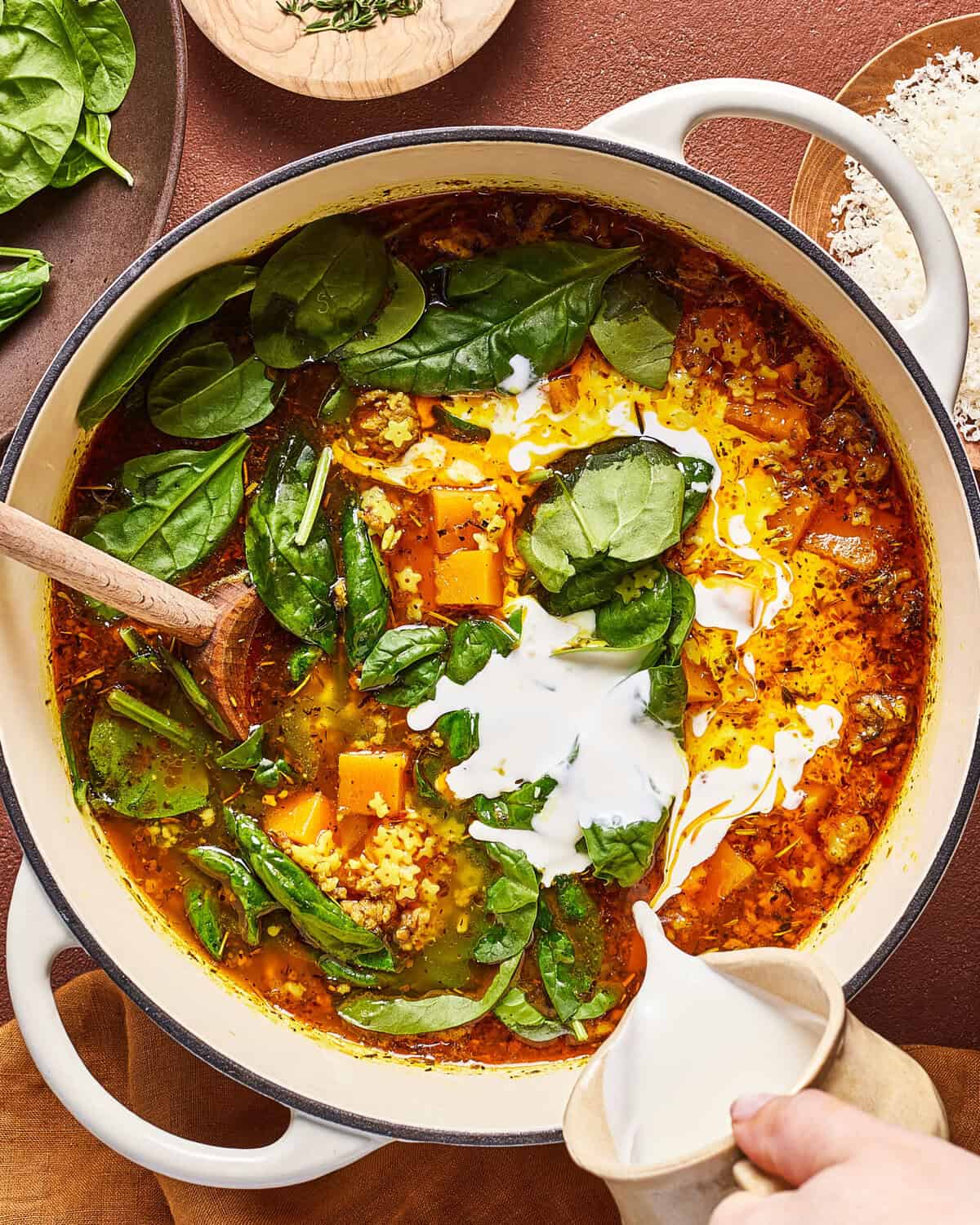 A hand pours cream into a pot of soup containing spinach leaves, cubes of butternut squash, grains, and spices. A wooden spoon rests in the pot, and fresh spinach and grated cheese are nearby.