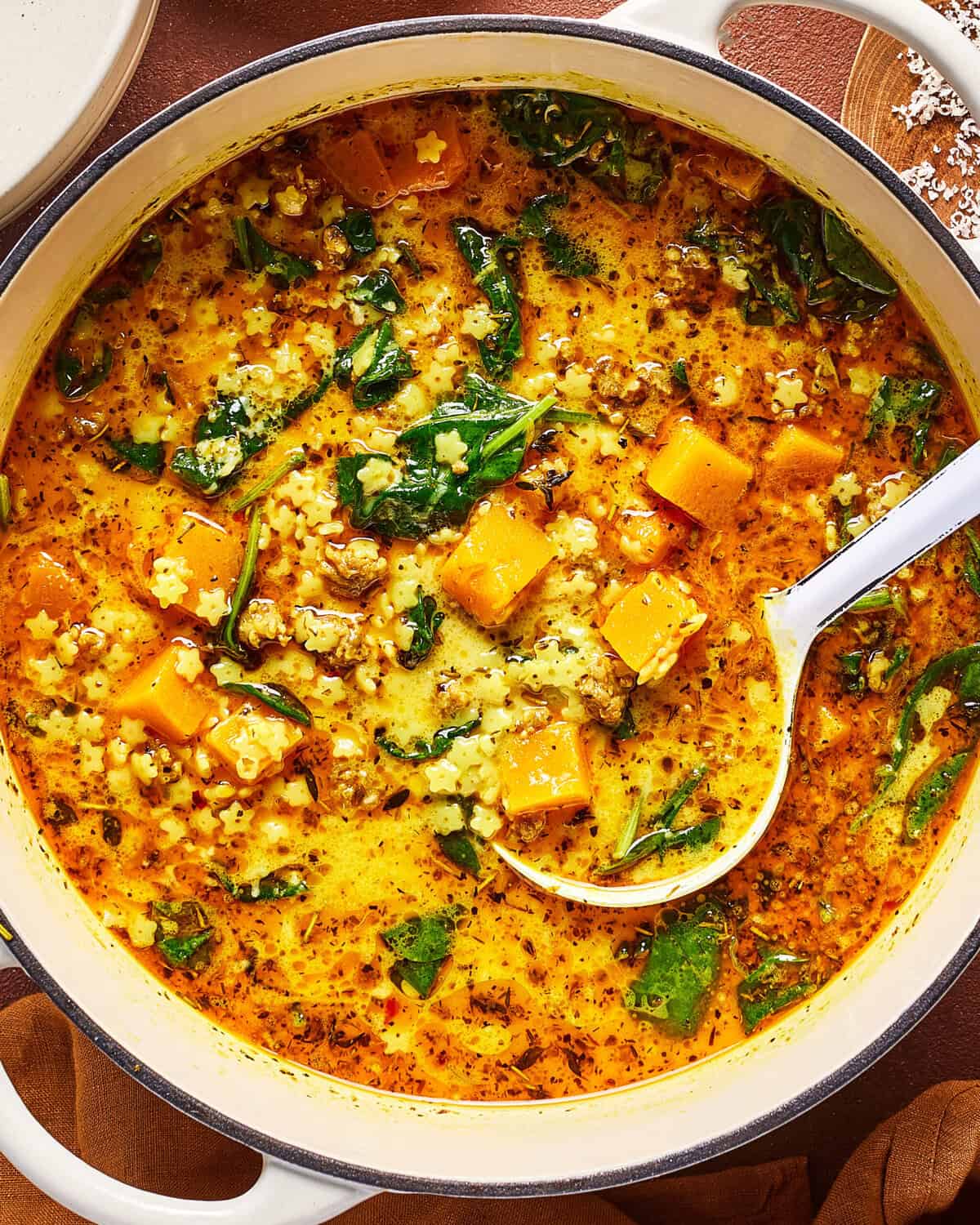 A pot of soup with chunks of orange butternut squash, leafy greens, small pasta, and herbs in a creamy, golden broth. A white ladle rests inside the pot.