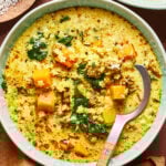 A bowl of creamy soup with yellow broth, chunks of butternut squash, small pasta, wilted greens, herbs, and spices, served with a spoon. A bowl of grated cheese is partly visible in the background.