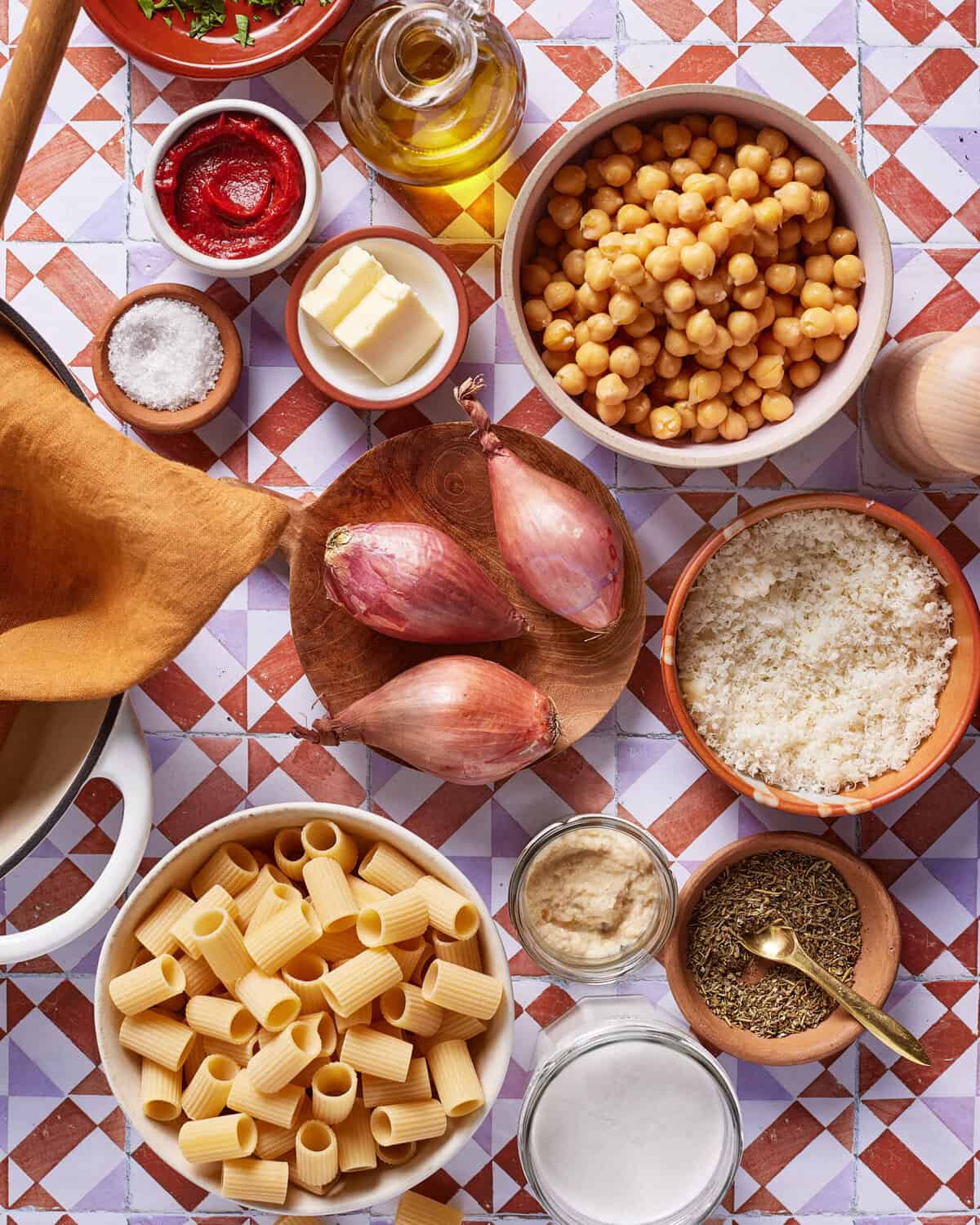Ingredients for cooking displayed on a patterned surface, including uncooked rigatoni pasta, chickpeas, shallots, tomato paste, grated cheese, butter, olive oil, seasonings, and salt.