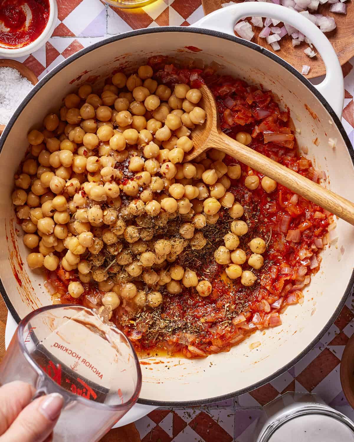 A hand pours water from a measuring cup into a pot filled with chickpeas, tomato sauce, onions, and spices. A wooden spoon rests in the pot, and various ingredients are visible around it on a tiled surface.
