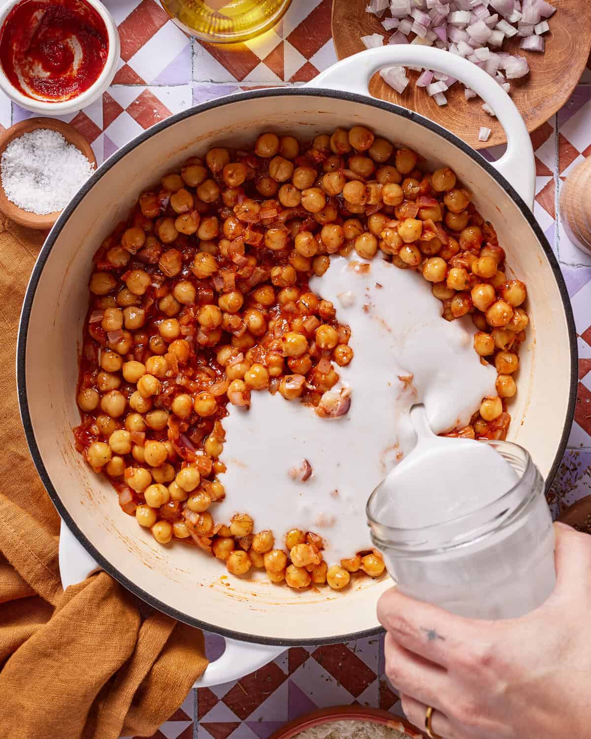 A hand pours coconut milk from a jar into a pot of chickpeas, tomato sauce, and onions. Chopped onions, salt, tomato paste, and oil are arranged around the white pot on a patterned surface.
