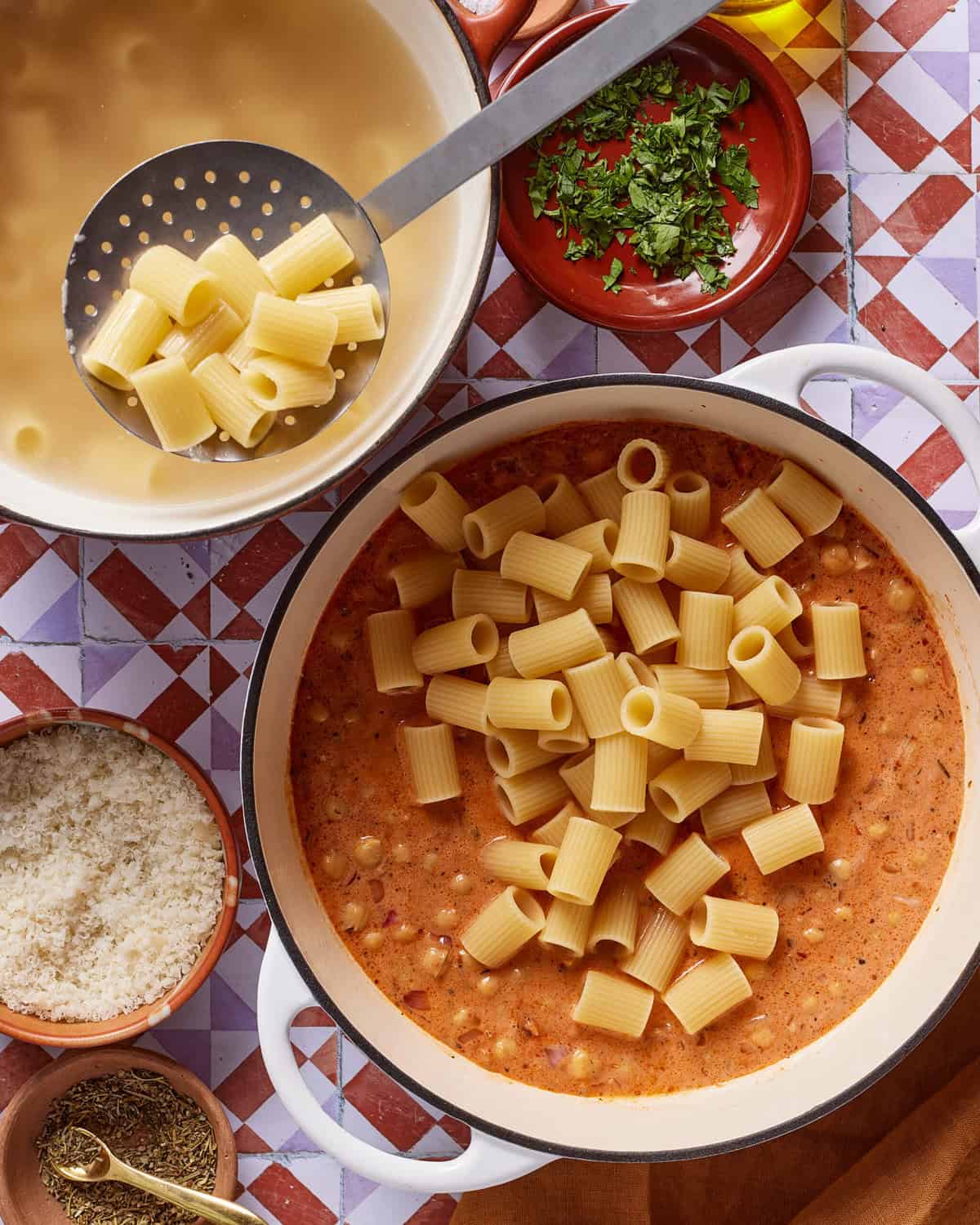 Overhead view of cooked rigatoni being added to a pot of creamy tomato sauce, surrounded by bowls of chopped herbs, grated cheese, and dried spices on a colorful tiled surface.