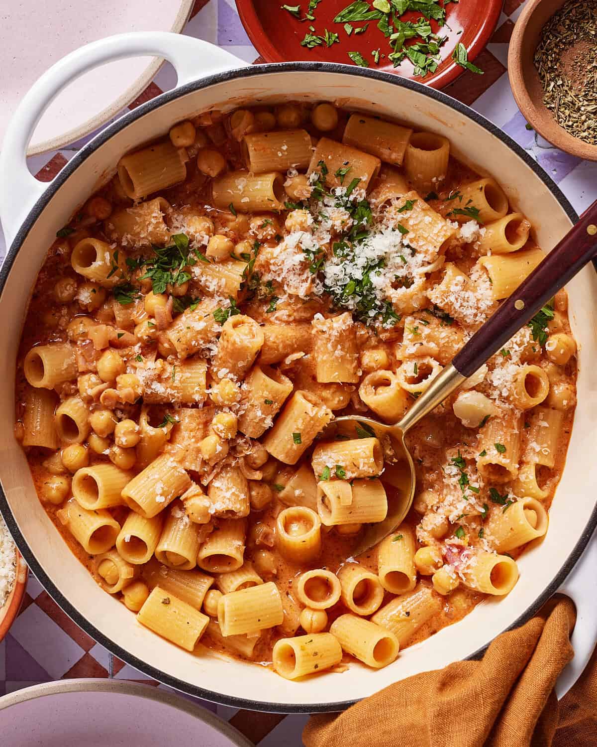A large white pot filled with creamy rigatoni pasta, chickpeas, tomato sauce, and topped with grated cheese and chopped parsley. A ladle rests in the pot, ready to serve. Plates and herbs surround the pot.