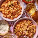 Two bowls of creamy tomato pasta with rigatoni and chickpeas, garnished with herbs. Surrounding the bowls are a glass of white wine, grated cheese, a small dish of herbs, and plates on a patterned table.
