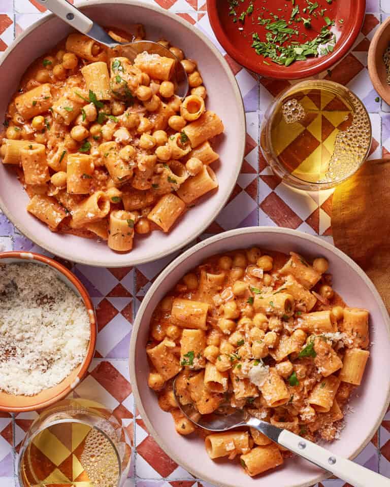 Two bowls of creamy tomato pasta with rigatoni and chickpeas, garnished with herbs. Surrounding the bowls are a glass of white wine, grated cheese, a small dish of herbs, and plates on a patterned table.