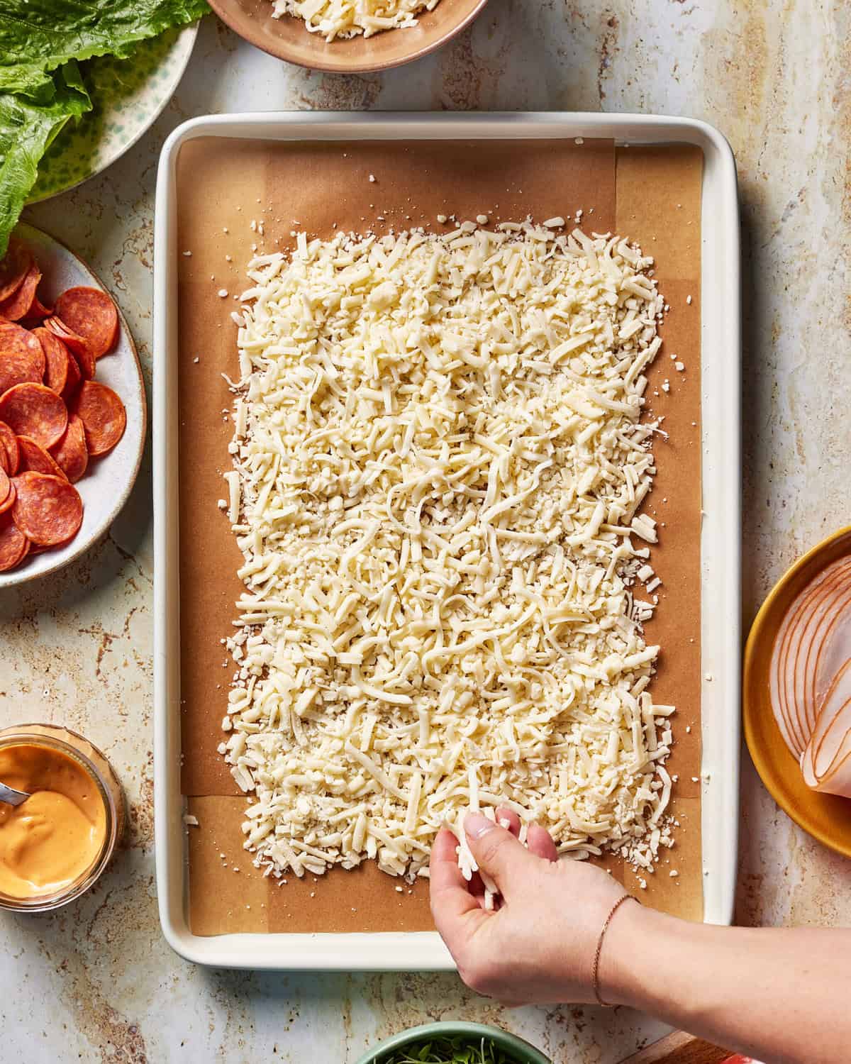A hand sprinkles shredded cheese over a rectangular baking sheet lined with parchment paper. Surrounding the sheet are plates of pepperoni, deli meat, lettuce, a bowl of sauce, and greens on a light countertop.
