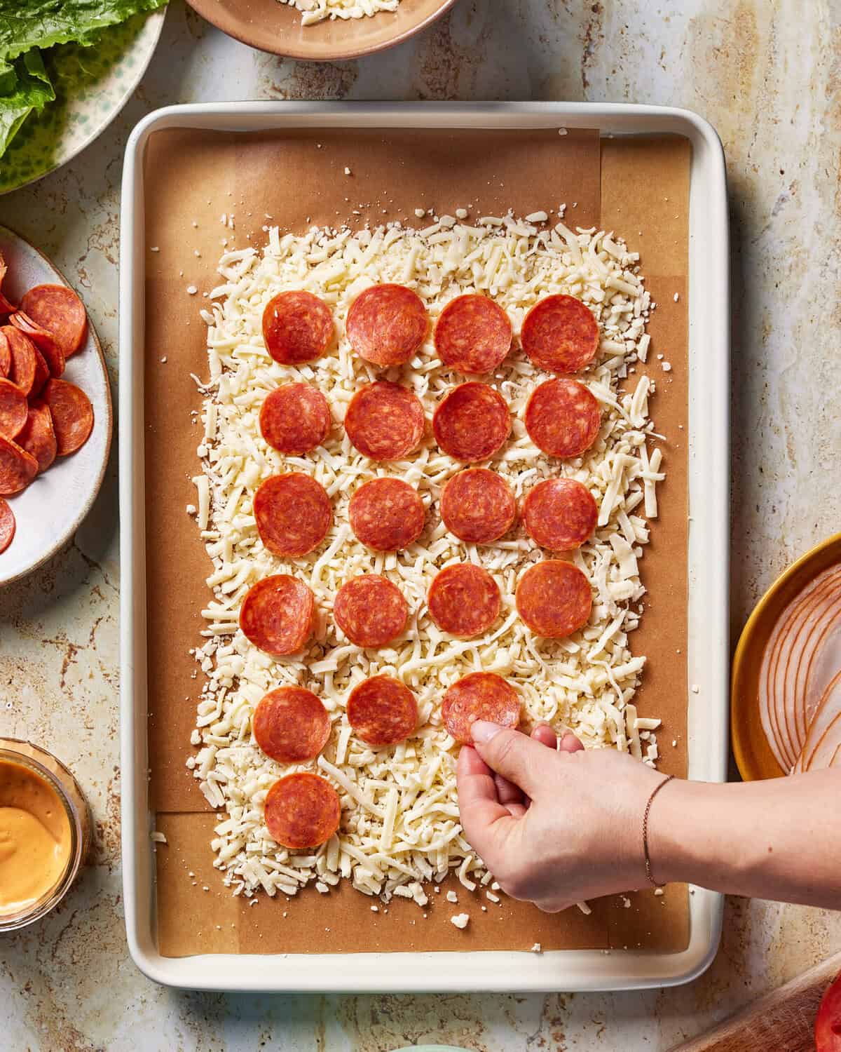A hand places pepperoni slices on a rectangular sheet of dough covered with shredded cheese, set on a parchment-lined baking sheet. Bowls of cheese, pepperoni, sauce, and greens are nearby.