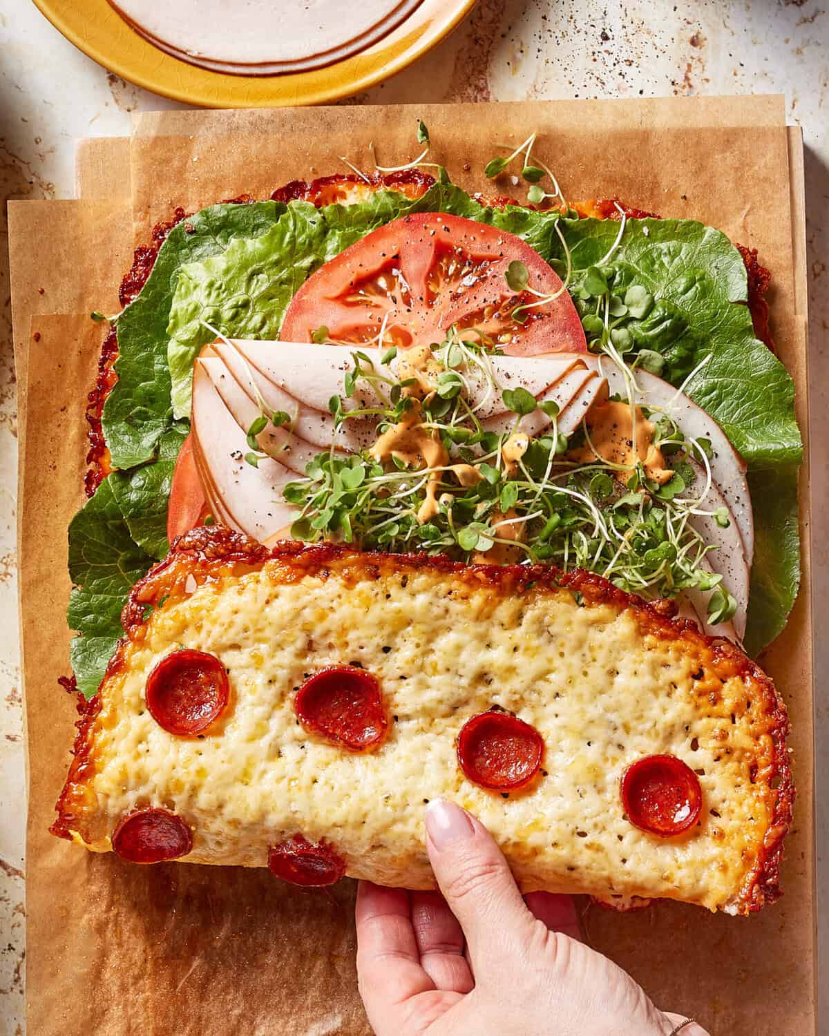 A hand folds a piece of cheesy pepperoni crust over a sandwich filled with lettuce, tomato, turkey slices, and microgreens, set on parchment paper atop a cutting board.