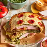 A crunchy pepperoni wrap with leafy greens, turkey, tomato slices, and cheese, stacked between two slices of bread topped with melted cheese and mini pepperoni. Nearby are a bowl of greens, sliced tomato, sauce, and deli meat.