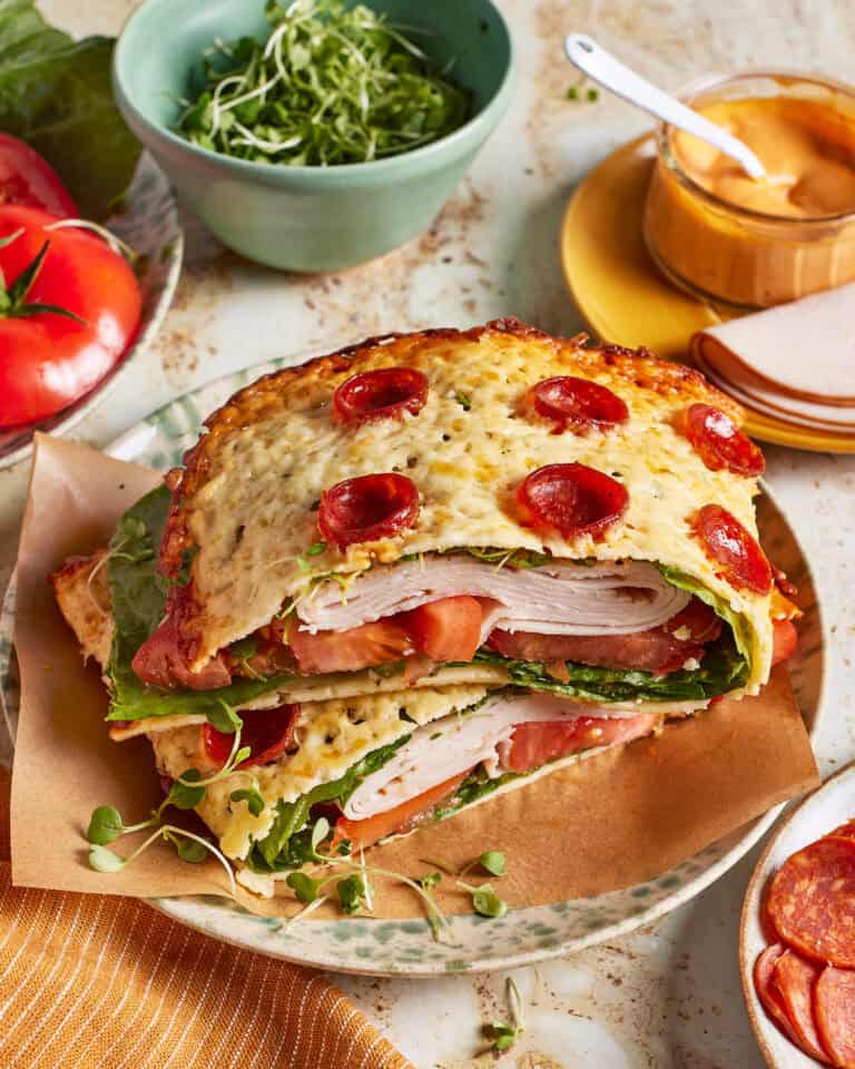 A crunchy pepperoni wrap with leafy greens, turkey, tomato slices, and cheese, stacked between two slices of bread topped with melted cheese and mini pepperoni. Nearby are a bowl of greens, sliced tomato, sauce, and deli meat.
