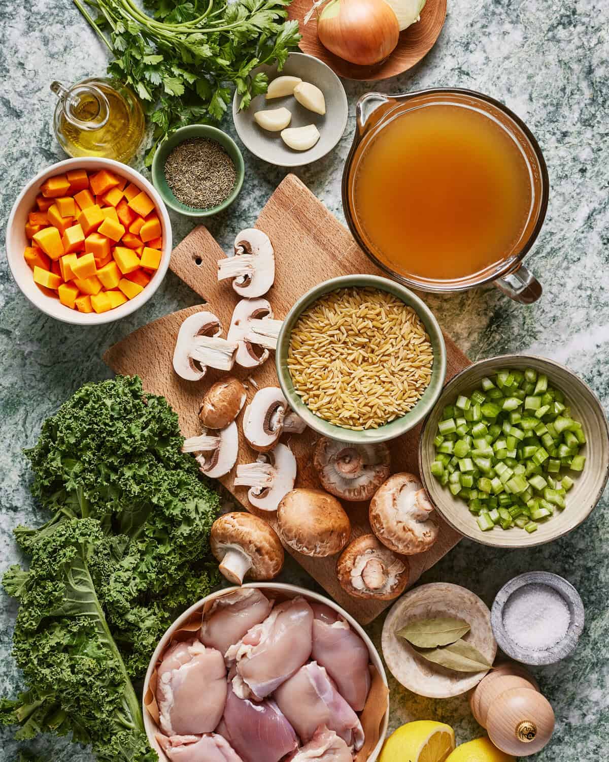 Assorted soup ingredients on a countertop, including chopped carrots, celery, mushrooms, kale, raw chicken pieces, dry rice, broth, garlic, onion, herbs, oil, salt, pepper, and a lemon.