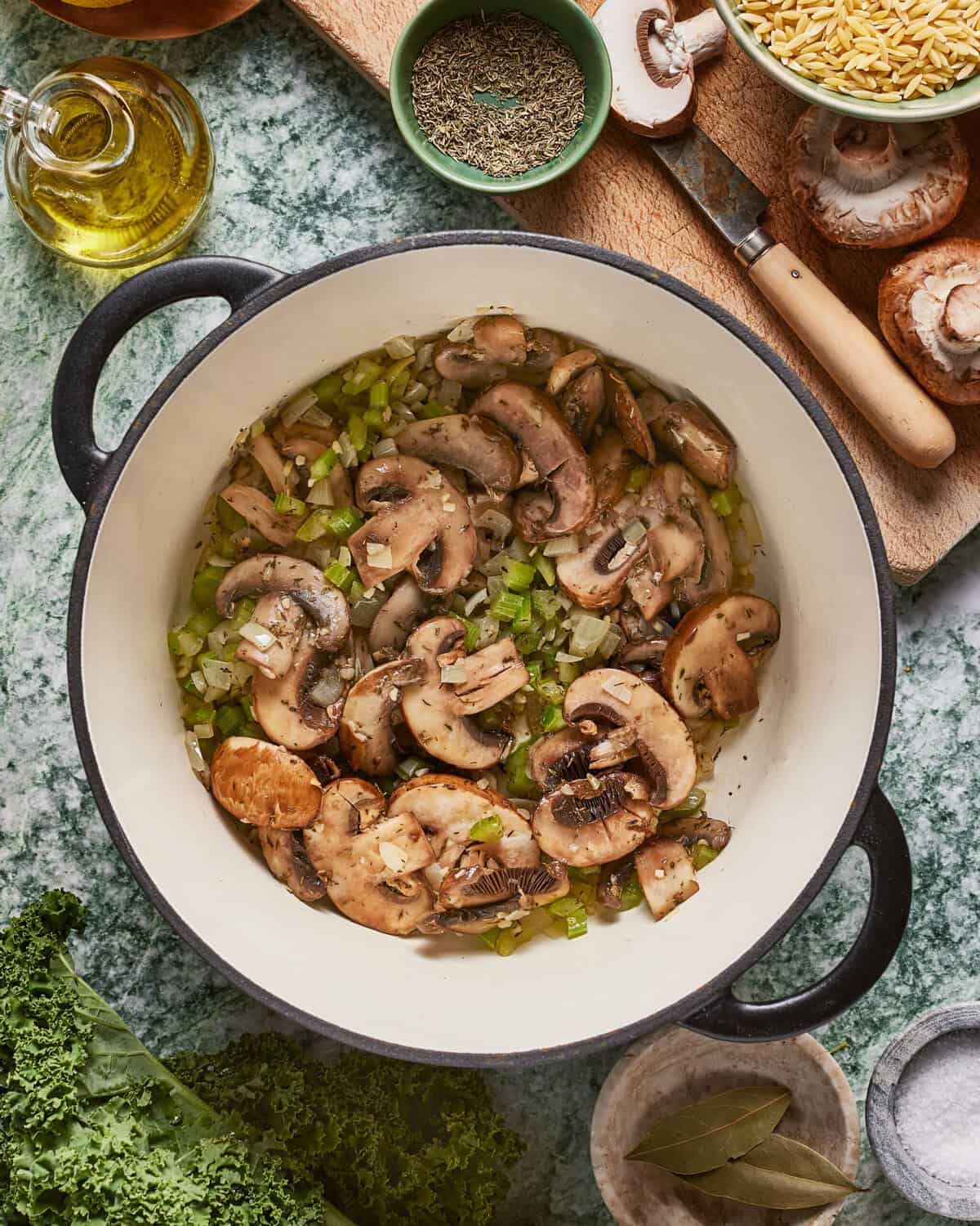 A white pot filled with sautéed mushrooms, celery, and onions sits on a green countertop. Surrounding the pot are fresh kale, mushrooms, orzo pasta, bay leaves, and a small bowl of dried herbs.