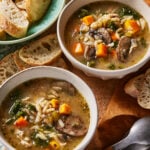 Two bowls of vegetable and sausage soup with orzo pasta, carrots, celery, mushrooms, and greens, served with slices of crusty bread on a wooden surface. Spoons and more bread are nearby.