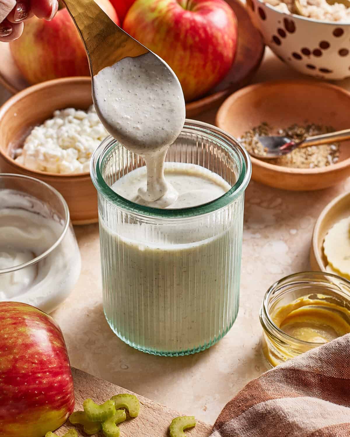 A glass jar filled with a creamy dressing is being stirred by a spoon. Surrounding the jar are apples, cottage cheese, yogurt, mustard, spices, and celery, arranged on a light kitchen countertop.