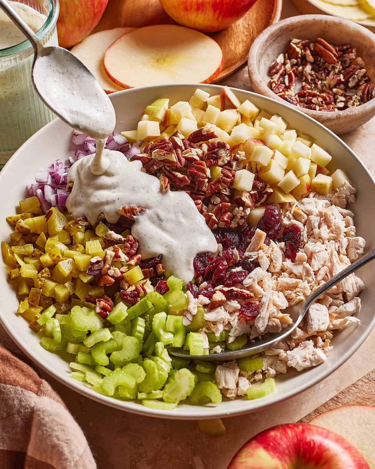 A bowl filled with chopped celery, pickles, red onion, apple, shredded chicken, dried cranberries, and pecans, with creamy dressing being spooned on top. A spoon and fork rest in the bowl, with apples and pecans nearby.