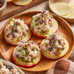 Four round apple slices topped with cheese and a chunky chicken salad mixture containing celery, red onion, and nuts, served on a wooden plate with sliced apples and cheese in the background.