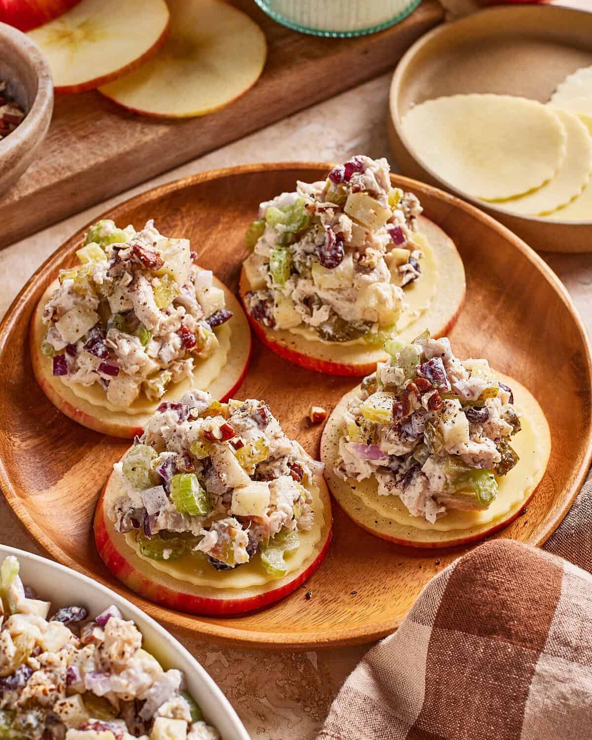 Four round apple slices topped with cheese and a chunky chicken salad mixture containing celery, red onion, and nuts, served on a wooden plate with sliced apples and cheese in the background.