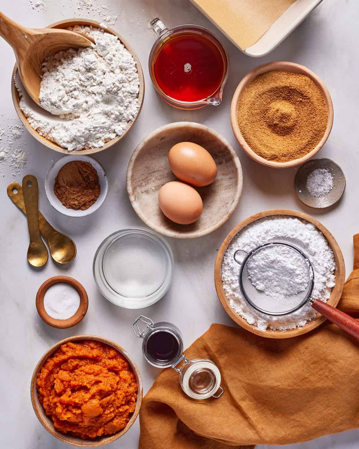 A flat lay of baking ingredients on a white surface, including flour, eggs, brown sugar, powdered sugar, cinnamon, baking powder, pumpkin puree, vanilla, oil, maple syrup, and measuring spoons.