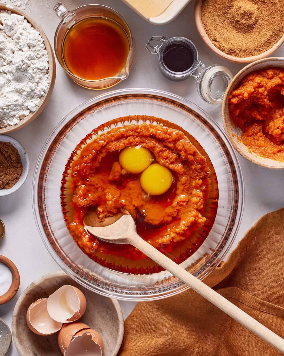 A glass bowl with pumpkin puree and two eggs being mixed with a wooden spoon, surrounded by bowls of flour, sugar, spices, vanilla, oil, and eggshells on a light countertop.