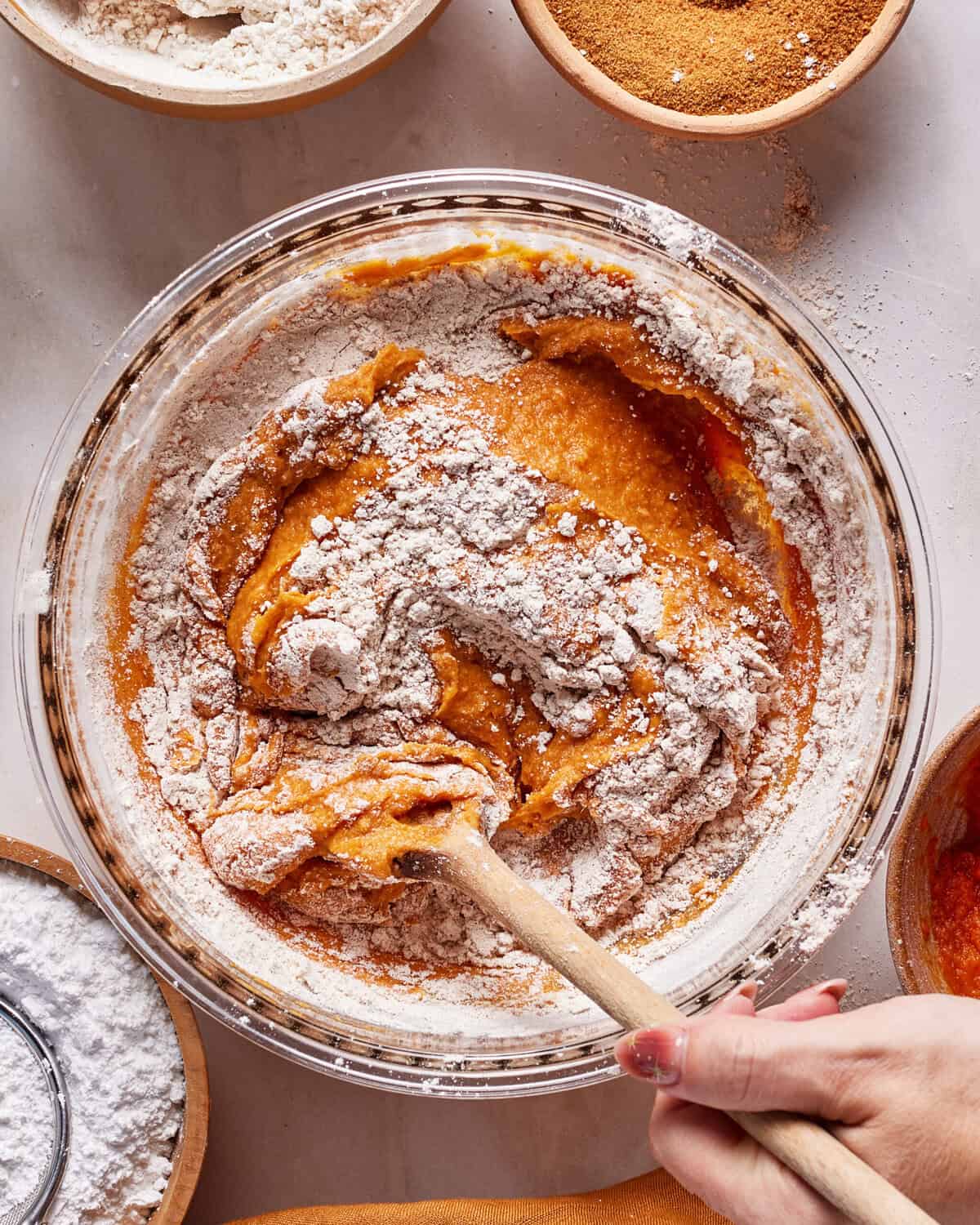 A hand stirs together flour and a pumpkin-colored batter in a glass bowl with a wooden spoon, surrounded by bowls of flour and spices on a white countertop.