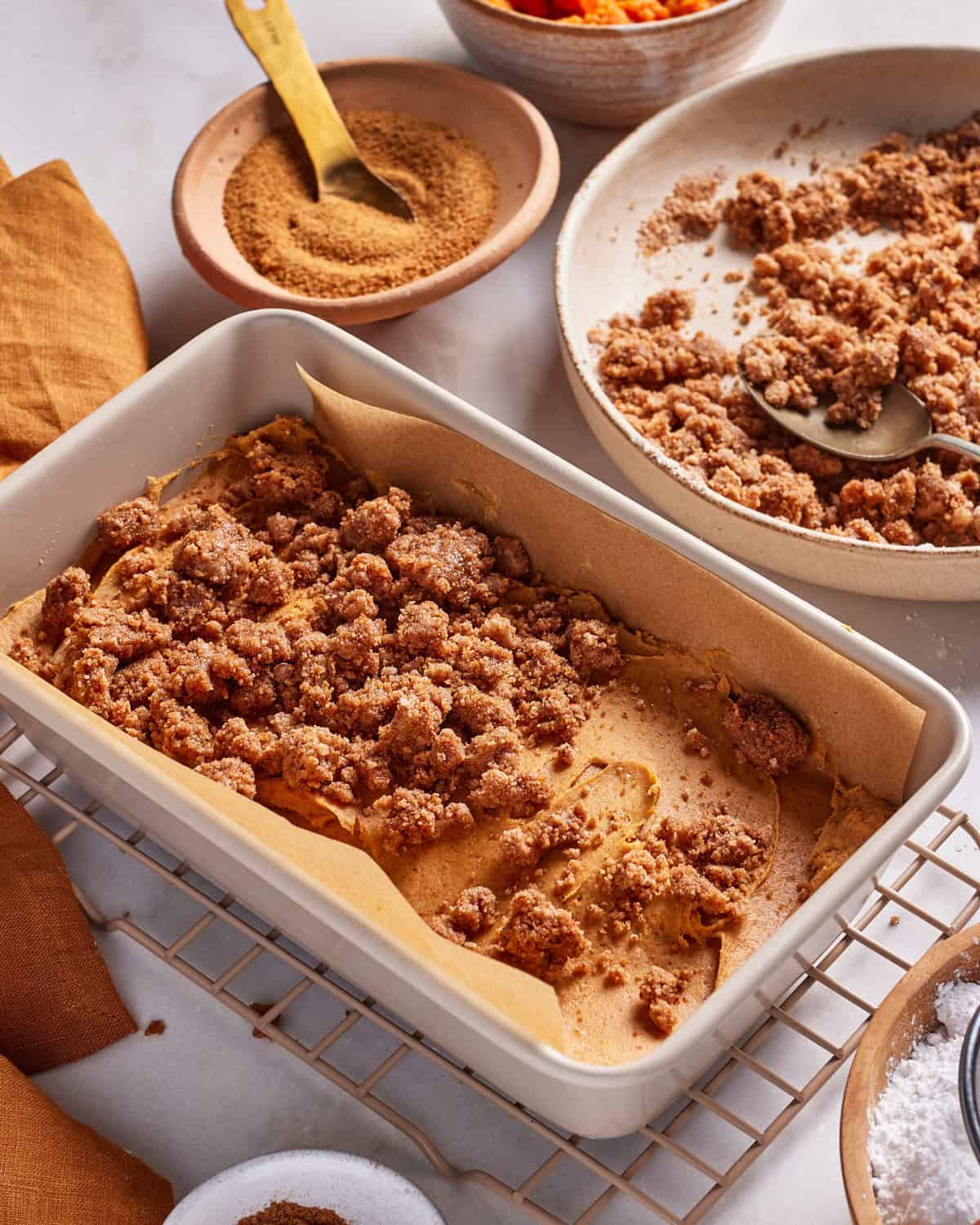 Rectangular baking dish lined with parchment paper and filled with a pumpkin dessert topped with brown sugar crumble. Bowls of streusel topping, brown sugar, and powdered sugar surround the dish on a cooling rack.