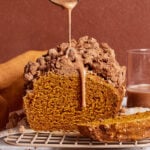 A loaf of pumpkin bread with a crumb topping sits on a cooling rack. A spoon drizzles icing over the top. A slice is cut in front, and a glass with brown liquid is in the background. The scene has warm brown tones.