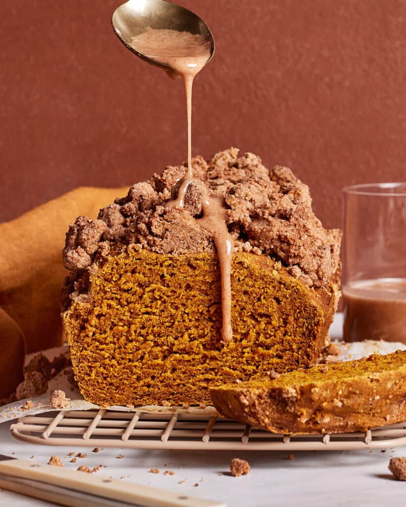 A loaf of pumpkin bread with a crumb topping sits on a cooling rack. A spoon drizzles icing over the top. A slice is cut in front, and a glass with brown liquid is in the background. The scene has warm brown tones.
