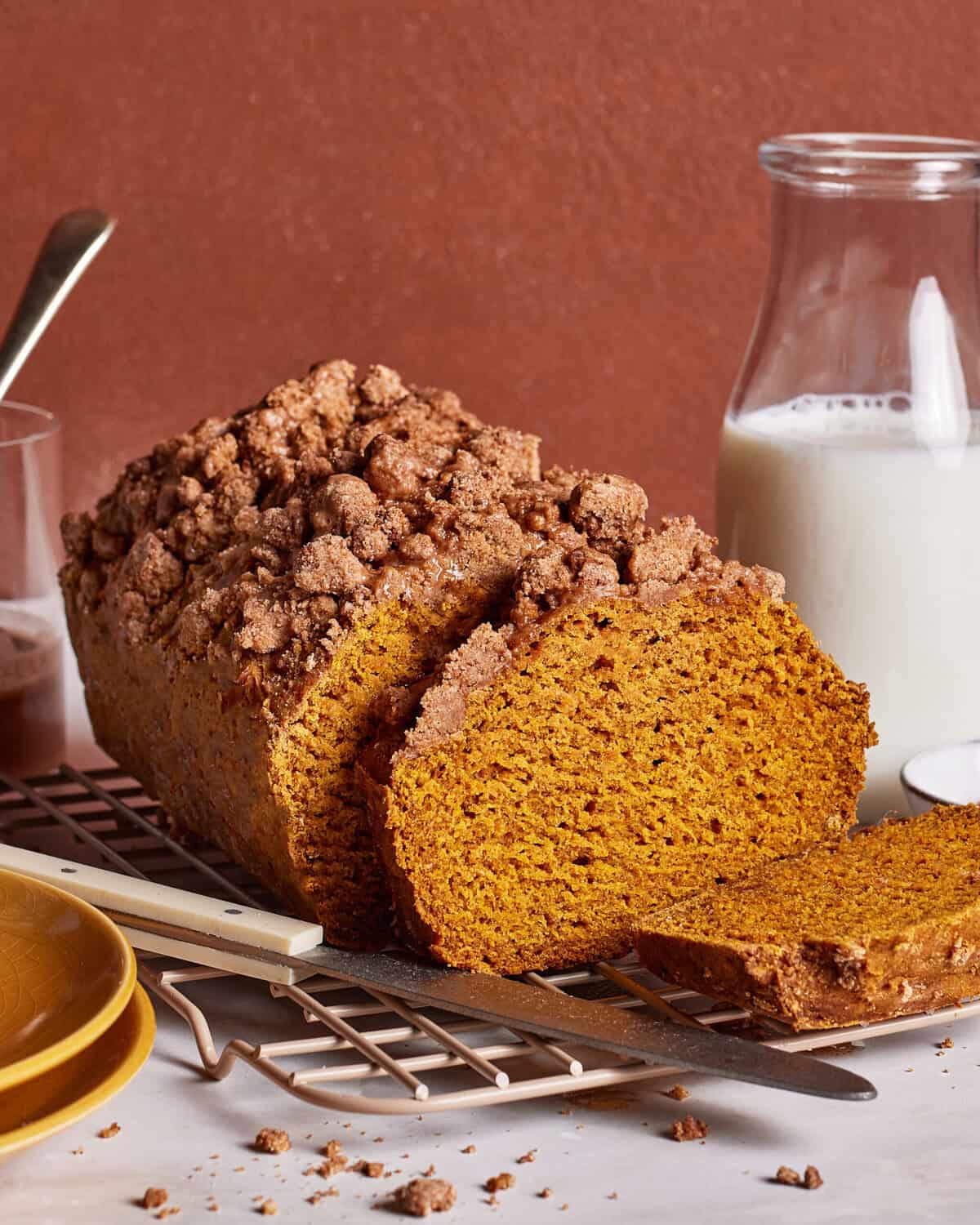 A loaf of pumpkin bread with a crumb topping sits on a cooling rack, partially sliced. A knife, plates, and a glass jug of milk are nearby on a white surface against a brown background.