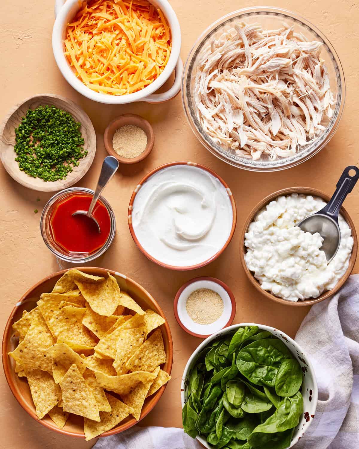 A variety of ingredients in bowls on a light surface, including shredded cheddar cheese, shredded chicken, chopped chives, hot sauce, sour cream, cottage cheese, tortilla chips, spinach leaves, and seasonings.