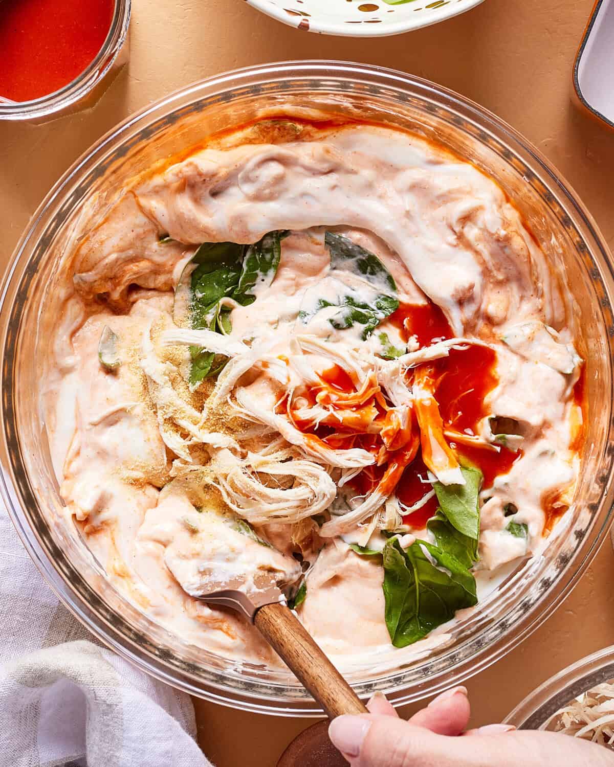 A glass bowl filled with a creamy mixture, shredded chicken, fresh spinach leaves, shredded cheese, and hot sauce being stirred with a wooden spoon. Ingredients are partially blended.