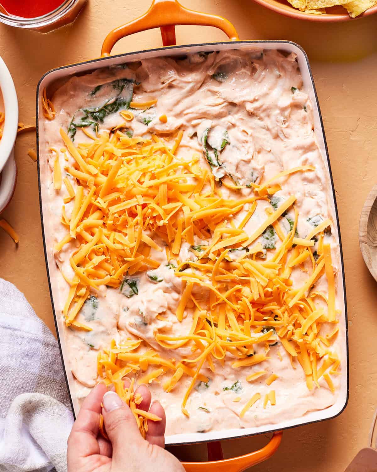 A hand sprinkles shredded cheddar cheese onto a creamy spinach dip mixture in a rectangular baking dish, preparing it for baking. Surrounding the dish are plates and a bowl of chips.