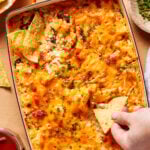 A hand dips a tortilla chip into a cheesy, high protein buffalo chicken dip topped with chopped chives in a rectangular orange baking dish. More tortilla chips are on the side.