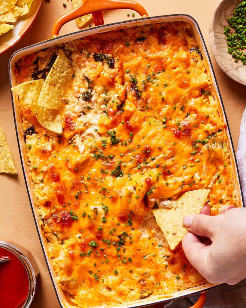 A hand dips a tortilla chip into a cheesy, high protein buffalo chicken dip topped with chopped chives in a rectangular orange baking dish. More tortilla chips are on the side.
