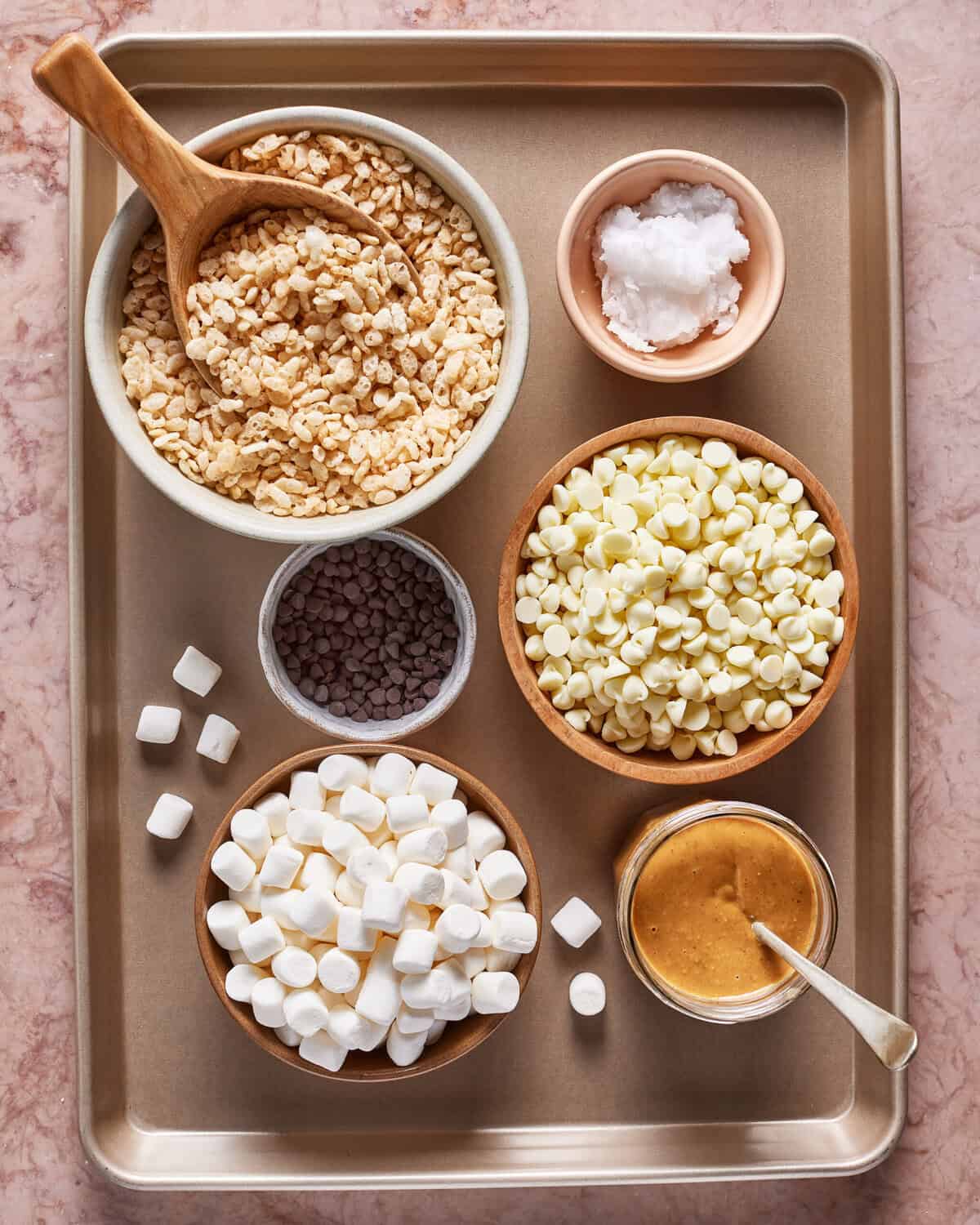 A baking tray holds bowls of rice cereal, mini marshmallows, white chocolate chips, peanut butter, coconut oil, and chocolate chips, with some loose marshmallows scattered nearby.