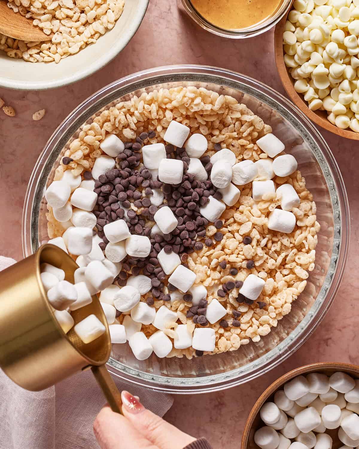 A hand pours mini marshmallows into a glass bowl filled with rice cereal, chocolate chips, and more marshmallows. Surrounding bowls hold rice cereal, peanut butter, and white chocolate chips.