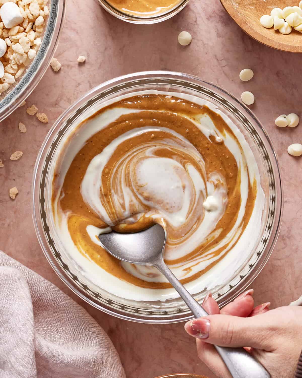 A hand holds a spoon in a glass bowl filled with swirled peanut butter and yogurt, surrounded by white chocolate chips, puffed rice, and a light pink cloth on a pink surface.