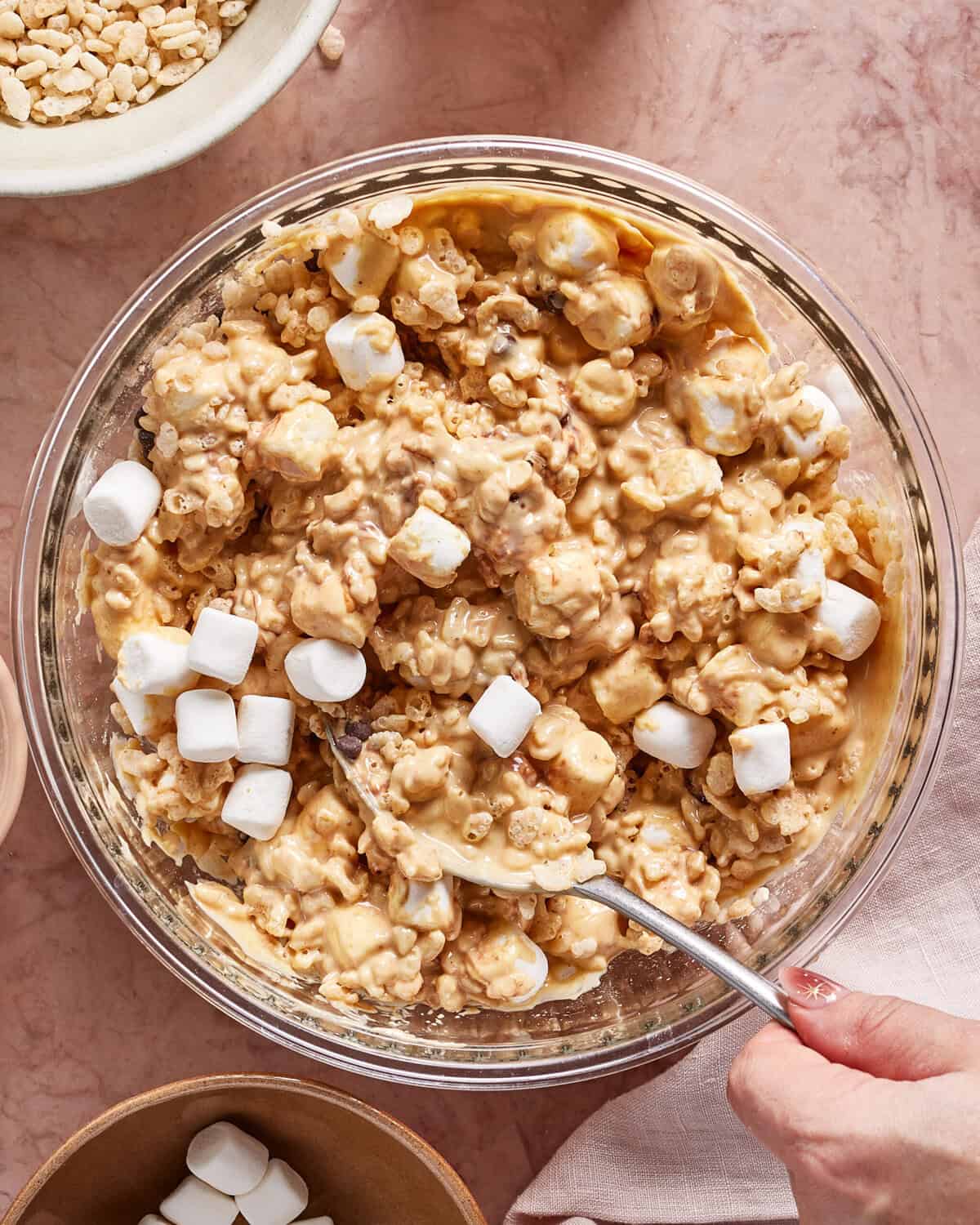 A hand stirs together Rice Krispies, peanut butter, and mini marshmallows in a glass mixing bowl on a pink countertop. Some unmixed marshmallows are visible on top of the mixture.