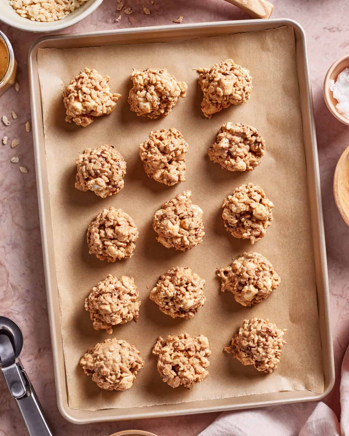 A baking sheet lined with parchment paper holds fifteen evenly spaced, unbaked cookie dough balls made with oats and rice cereal. Mixing bowls and a scoop are visible around the tray on a pink countertop.