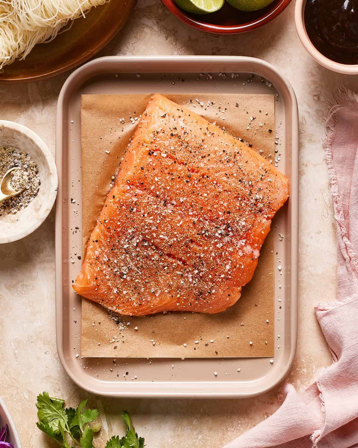 A raw salmon fillet seasoned with salt and pepper sits on brown parchment paper in a beige tray, surrounded by various ingredients including herbs, lime halves, spices, and noodles on a light-colored surface.