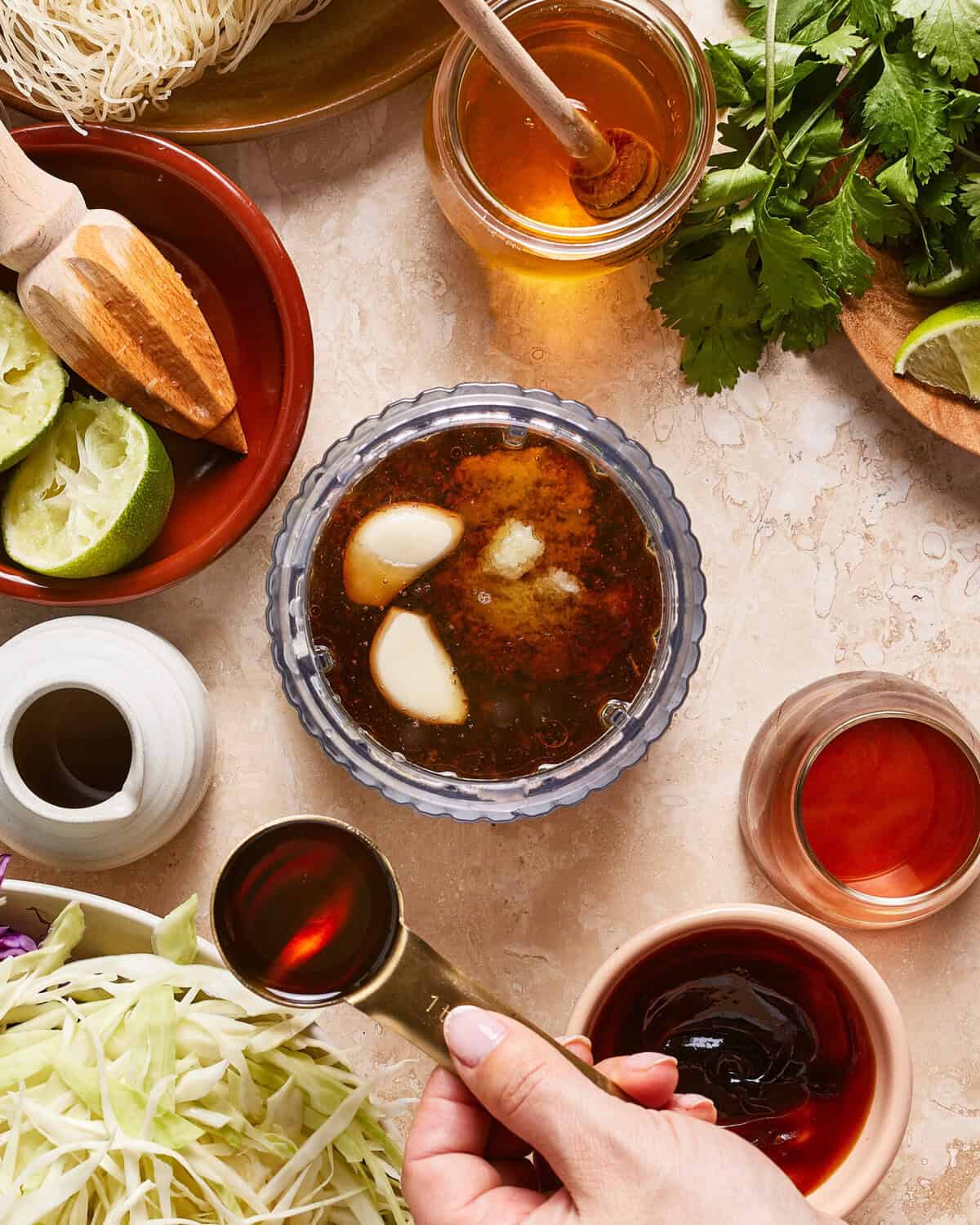 A hand pours a dark liquid into a bowl of sauce with garlic cloves, surrounded by limes, honey, cabbage, herbs, and various small jars and bowls of ingredients on a light countertop.